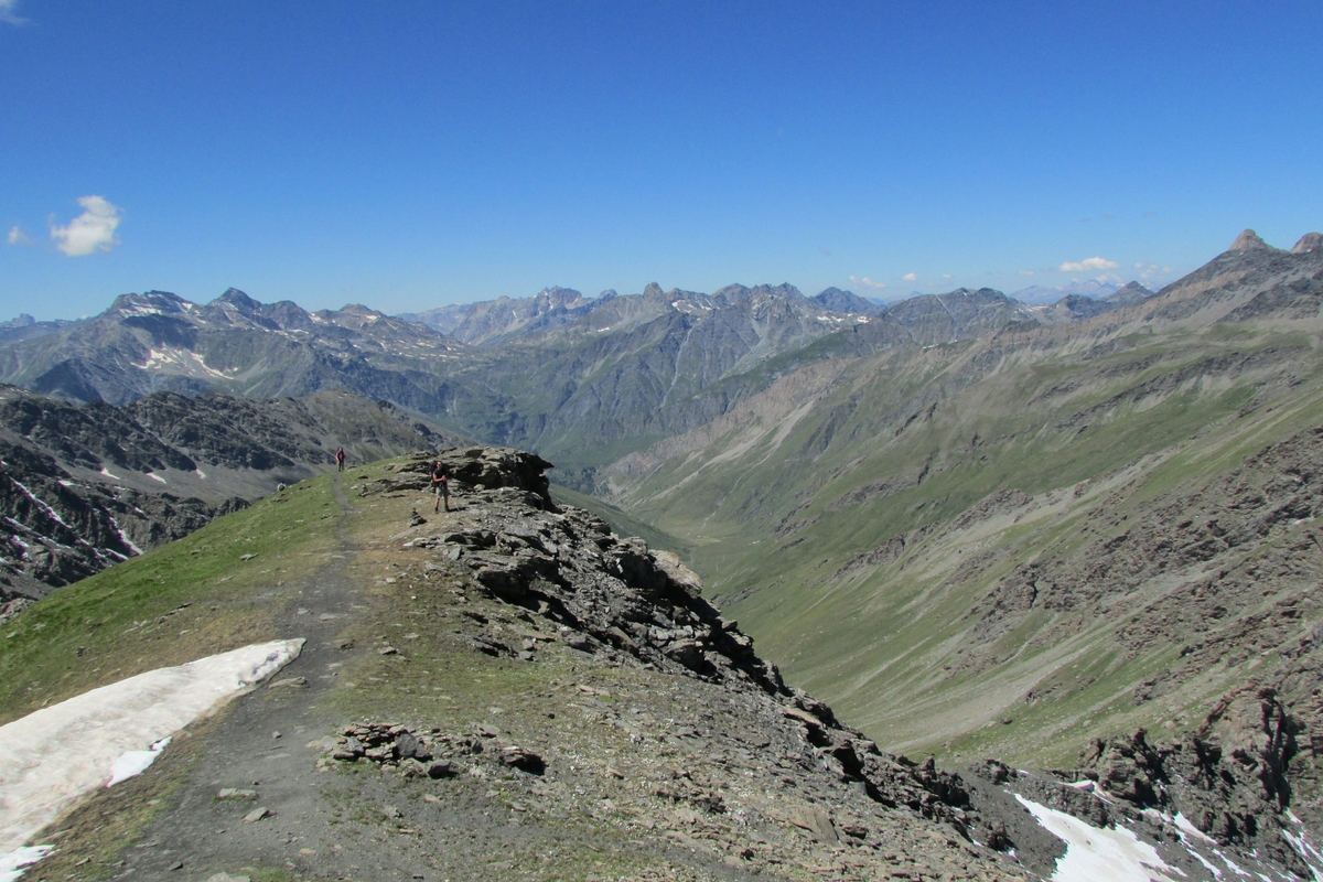 pianoro in alta quota con lembo di neve, rocce e radi prati, che si innalza su vallone sottostante. Due persone in secondo piano. intorno la catena montuosa