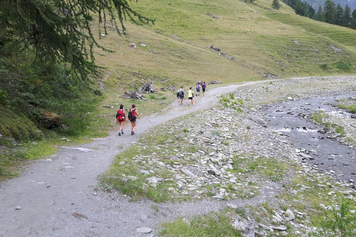Su un sentiero lungo un piccolo corso d'acqua ci sono alcuni escursionisti di schiena. Due ragazze in primo piano con zaino rosso e quattro ragazzi più avanti. intorno pendii erbosi e qualche larice